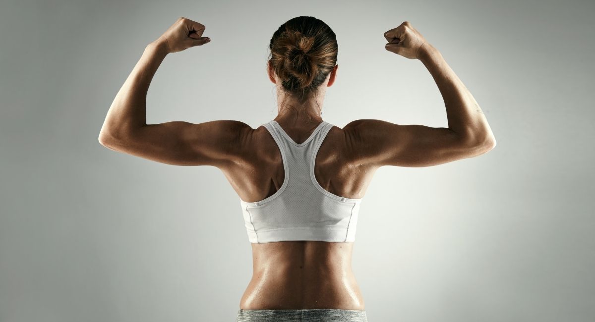 A fit woman in a white sports bra viewed from behind flexing both arms to show her muscular back, representing strength, discipline, and the life-changing habits needed to build rock-solid self-confidence from scratch.