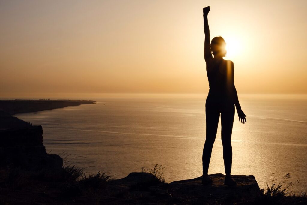 A confident woman in the mountain raising her left arm facing sunset.